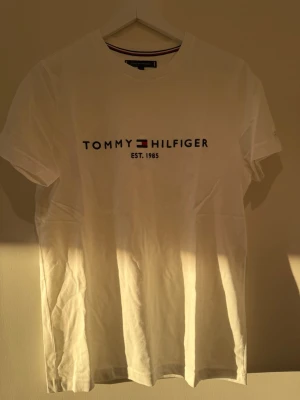Vit t-shirt från Tommy Hilfiger - Vit t-shirt från Tommy Hilfiger med klassisk logotyp och text 'Est. 1985' på bröstet. T-shirten har rund hals, korta ärmar och är tillverkad i mjuk bomull. Perfekt för dig som gillar stilrena och ikoniska plagg.
