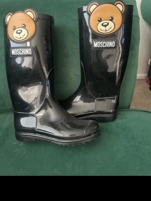 Svarta gummistövlar Moschino björn - Säljer ett par svarta gummistövlar från Moschino med den ikoniska björndesignen framtill. Stövlarna har hög skaft och tydlig logga på framsidan. Tillverkade i glansigt gummimaterial och perfekta för regniga dagar.