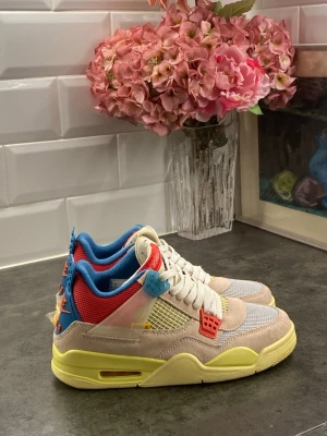Union LA x Air Jordan 4 Retro sneakers - Färgglada Union LA x Air Jordan 4 Retro sneakers med mesh och mocka i beige, grått och gult. Skorna har röda och blå detaljer, vit snörning och en gul sula. Kombinationen av material och färger ger en unik och trendig look.