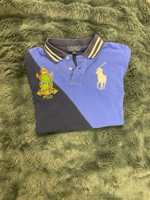 Blå och svart piké från Polo Ralph Lauren - Snygg pikétröja från Polo Ralph Lauren i blått och svart med broderad logga och emblem på bröstet. Klassisk krage med ränder och knappar framtill. Tillverkad i bomull för en bekväm känsla. Perfekt för en sportig och stilren look. Storlek M 10-12