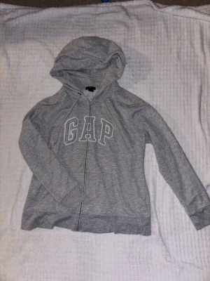 Grå zip hoodie från GAP - Snygg gråmelerad hoodie från GAP med dragkedja framtill och stor logga broderad över bröstet. Tröjan har huva med dragsko och ribbade muddar vid ärmslut och nederkant. Obs liten i storleken, mer som en M/L