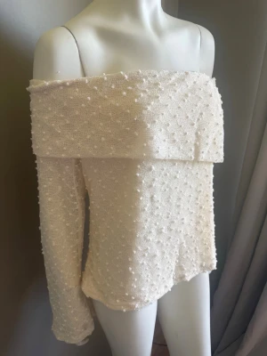 Vit offshoulder topp med struktur strl XL - Superfin vit offshoulder topp med lång transparent ärm och markerad volangkant upptill. Toppens tyg har en strukturerad, prickig yta som ger en unik look. Perfekt för dig som vill sticka ut med en trendig och elegant stil.