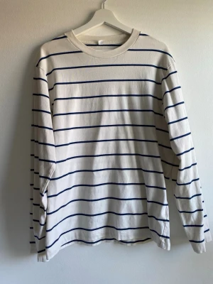 Randig långärmad tröja från Uniqlo - Vit långärmad tröja från Uniqlo med tunna mörkblå horisontella ränder. Tillverkad i 100% bomull, har rund halsringning och avslappnad passform. Perfekt för en stilren och enkel look.