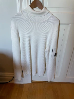 Vit turtleneck  från C.P. Company - Säljer en stilren vit turtleneck från C.P. Company med ribbstickad struktur och lång ärm. Tröjan har en hög krage och en ikonisk linsdetalj på ena ärmen. Perfekt för lager-på-lager, användes typ 1 gång på nyårsafton 