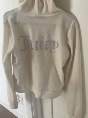 Vit Juicy Couture hoodie med strass - Snygg vit hoodie från Juicy Couture med dragkedja och huva. Baksidan har texten 'Juicy' i glittrande strass och framsidan har en liten logga. Mjuk velourkänsla och kängurufickor framtill. Perfekt för en trendig och avslappnad look.