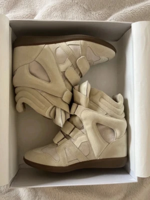 Isabel Marant beige höga sneakers - Säljer ett par ikoniska höga sneakers från Isabel Marant i beige mocka och textil. Skorna har flera kardborreband, coola paneldetaljer och en chunky sula i brunt gummi. Perfekta för dig som vill ha en statement sneaker med unik design.