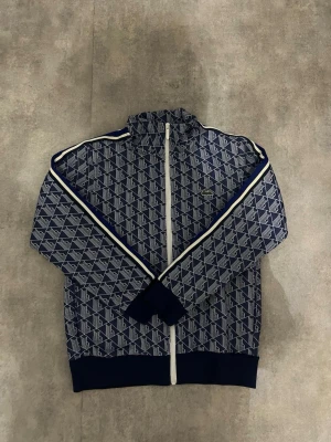Blå Lacoste hoodie med mönster - Snygg blå hoodie från Lacoste med hel dragkedja och vit logotypmönster över hela plagget. Huva och muddar i mörkblått, samt vita ränder längs ärmarna. Perfekt för en sportig och trendig look.