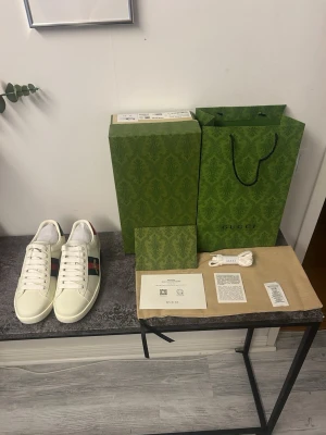 Gucci vita sneakers med röd/grön rand - Snygga vita sneakers från Gucci i läder med klassiska röd och grön rand på sidan samt diskret logga på plösen. Skorna har vita snören och en stilren låg silhuett. Perfekta för dig som vill ha en exklusiv touch på din outfit.