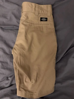 Beige shorts från Dickies - Säljer ett par klassiska beige shorts från Dickies med hög midja och en enkel design. Shortsens material är slitstark bomull och de har en bakficka med Dickies-logga samt dragkedja framtill. Perfekta för dig som gillar streetwear och enkel stil.