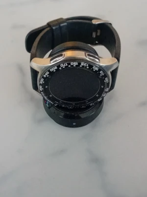 Samsung smartklocka med silikonarmband - Snygg svart smartklocka med rund urtavla och svart silikonarmband. Klockan har en silverfärgad ram och markerad bezel med siffror. Perfekt för dig som vill ha koll på både tid och notiser direkt på handleden. Laddare saknas och klockan startar ej.