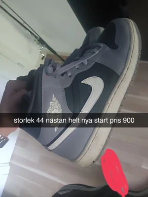 Nike Air Jordan 1 Mid grå/svart - Snygga Nike Air Jordan 1 Mid sneakers i grått och svart med vit swoosh och klassisk Air Jordan-logga på sidan. Skorna har en hög siluett och är tillverkade i läder med matchande grå snörning och vit sula.