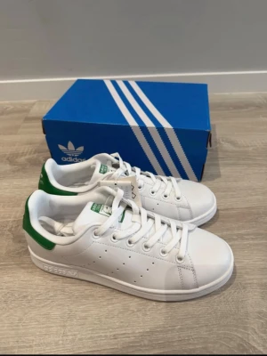 Adidas Stan Smith vita sneakers - Klassiska vita Adidas Stan Smith sneakers med gröna detaljer på hälen och tungan. Skorna har en låg siluett, perforerade ränder på sidorna och är tillverkade i slätt läder. Perfekta för en clean och tidlös stil. Helt nya!