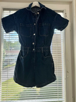 Mörkblå jeansjumpsuit med kort ärm - Snygg mörkblå jeansjumpsuit med korta ärmar och shortsben. Har två bröstfickor, två sidofickor och två bakfickor. Knäppning framtill med silverfärgade knappar och markerad midja med hällor för skärp. Perfekt för en avslappnad och trendig look. 