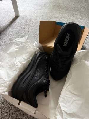 Svarta Hoka sneakers med chunky sula - Snygga helsvarta sneakers från Hoka med chunky sula och ventilerande mesh upptill. Skorna har klassisk snörning och diskret Hoka-logga på sidan. Perfekta för dig som gillar sportig stil och vill ha bekväma skor med modern look.