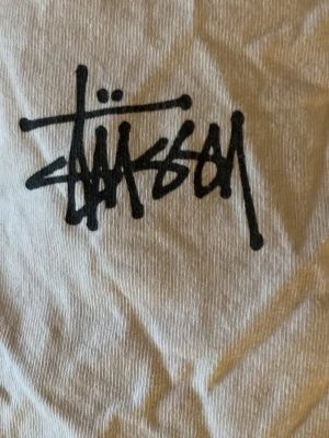 Vit Stüssy t-shirt med logga - Vit t-shirt från Stüssy med den ikoniska svarta loggan tryckt stort på ryggen. Klassisk passform och rund halsringning. Tillverkad i mjuk bomull som känns skön mot huden. Perfekt för dig som gillar streetwear och vill sticka ut.