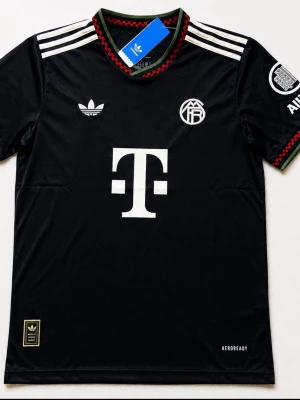 Svart Bayern München Adidas tröja - Snygg svart Bayern München matchtröja från Adidas med vita detaljer och klubbmärke på bröstet. Tröjan har korta ärmar, rund hals med röd och grön kant samt Adidas klassiska ränder på axlarna. Tillverkad i lätt och ventilerande material (AEROREADY).