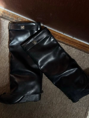 Svarta höga läderstövlar - Snygga svarta stövlar i läder med hög skaft och diskret metallogo upptill. Klassisk design med spetsig tå och liten klack. Perfekta för att lyfta din outfit med en stilren touch. Väldigt trasiga och slitna som det syns på bilderna, men kanske någon som gillar att laga skor eller något? Annars kastas dom.
