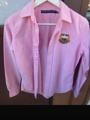Rosa skjorta från Ralph Lauren - Snygg rosa skjorta från Ralph Lauren med broderad logga på bröstet. Klassisk krage, knäppning framtill och lång ärm. Perfekt för dig som vill ha en stilren och preppy look.