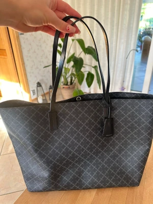 Svart axelväska By Malene Birger - Svart axelväska från By Malene Birger med klassiskt mönster i grått. Väskan har tunna axelband, stor öppen insida och en innerficka med dragkedja. Materialet är tåligt konstläder och detaljerna är i guld. Små skador längst ner vid fästena på ena sidan av väskan, där av såpass prissänkt. Annars knappt använd alls.