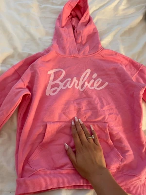 Rosa Barbie hoodie med huva - Cool rosa hoodie från Barbie med stor vit logga på bröstet. Tröjan har huva och en klassisk magficka framtill. Ribbstickade muddar vid ärmslut och nederkant. Perfekt för dig som gillar färg och ikoniska prints.