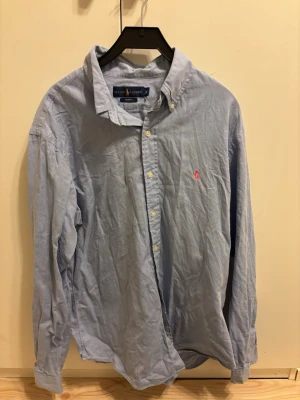 Ljusblå skjorta från Ralph Lauren - Klassisk ljusblå skjorta från Ralph Lauren med knappslå och krage. Skjortan har lång ärm och den ikoniska rosa logotypen broderad på bröstet. Tillverkad i mjuk bomull och perfekt för en clean och stilren look.