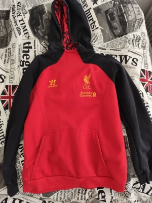 Liverpool FC röd och svart hoodie - Snygg röd och svart hoodie från Warrior med Liverpool FC-logga och Standard Chartered-tryck i gult på bröstet. Huva med mönstrat rött foder och känguruficka framtill. Perfekt för dig som älskar fotboll och vill ha en sportig stil.