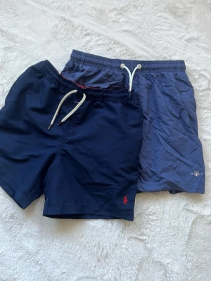  badbyxor Polo Ralph Lauren & Gant - Säljer två par snygga badbyxor, ett par mörkblå från Polo Ralph Lauren med vit dragsko och röd logga, samt ett par ljusblå från Gant med vit dragsko och diskret logga. Båda har klassisk passform och är tillverkade i snabbtorkande material.