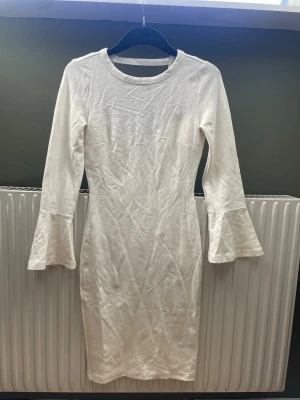 Vit långärmad klänning med trumpetärm - Säljer en enkel och stilren vit klänning med lång ärm och trumpetformade ärmslut. Klänningen har en kroppsnära passform och är gjord i mjuk bomull, perfekt för dig som kanske ska ta studenten. 🎓 