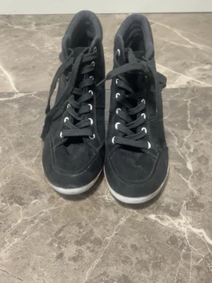 Svarta kilklackade sneakers - Snygga svarta sneakers med kilklack och snörning. Skorna har en hög siluett och är gjorda i mockaliknande material med vita detaljer på sulan. Perfekta för dig som vill ha lite extra höjd men ändå en sportig look.