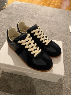 Maison margiella - Mörkblåa  sneakers med vita snören och brun sula. Skorna har en klassisk låg profil och är tillverkade i en mix av mocka och läder. Insidan är ljus och de har en stilren design som passar till det mesta. Helt nya, inte använda. 