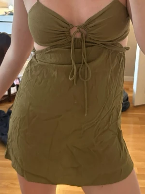 Olivgrön kort klänning med snörning - Säljer en olivgrön kort klänning med snörning framtill och cut-out detalj under bysten. Klänningen har smala axelband och är tillverkad i ett mjukt bomullsliknande material. Perfekt för varma dagar och ger en avslappnad men trendig look.