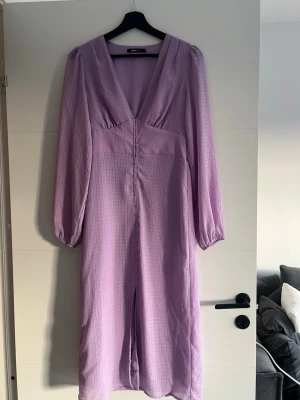 Ljuslila långklänning från Ginatricot - Säljer en ljuslila långklänning från Ginatricot med v-ringning, långa puffärmar och dekorativa knappar framtill. Klänningen har ett mjukt, strukturerat tyg och markerad midja för en snygg siluett. Perfekt för dig som vill ha en trendig och elegant look.