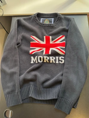  stickad tröja Morris - Snygg stickad tröja från Morris med Union Jack-flagga och texten 'MORRIS' framtill. Tröjan har ribbade muddar vid ärmslut och nederkant samt rund halsringning. Perfekt för dig som gillar klassisk brittisk stil.