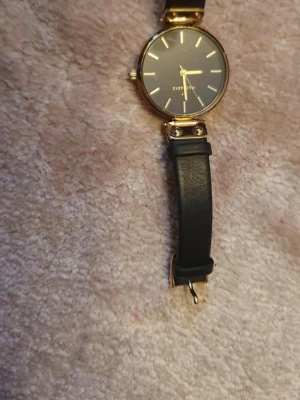 Svart och guld klocka från Mockaberg - Stilren klocka från  Mockaberg  med svart urtavla och guldfärgade detaljer. Klockan har ett klassiskt runt ur och svart armband i syntetmaterial. Perfekt accessoar för dig som gillar enkel men elegant design.