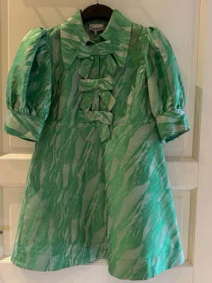 Grön kortklänning med rosetter Ganni - Supersöt grön kortklänning från Ganni med puffärmar, stora kragar och tre dekorativa rosetter framtill. Klänningen har ett unikt mönster i ljusgrönt och vitt och är tillverkad i ett glansigt material som känns lyxigt. Perfekt för dig som vill sticka ut!