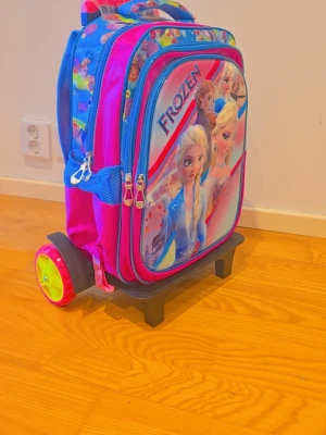 Frozen rullryggsäck med motiv - Säljer en färgglad rullryggsäck med motiv från Disney Frozen. Ryggsäcken har flera fack med dragkedja, meshfickor på sidorna och utdragbart handtag. Färgerna är blå, rosa och lila med tryck av Elsa och Anna. Perfekt för skolan eller resan!