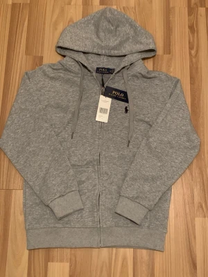 Grå hoodie från Polo Ralph Lauren - Snygg grå hoodie från Polo Ralph Lauren med dragkedja framtill och klassisk huva. Tröjan har ribbade muddar, fickor på sidorna och den ikoniska Polo-loggan broderad på bröstet. Tillverkad i mjukt bomullsmaterial för extra komfort.