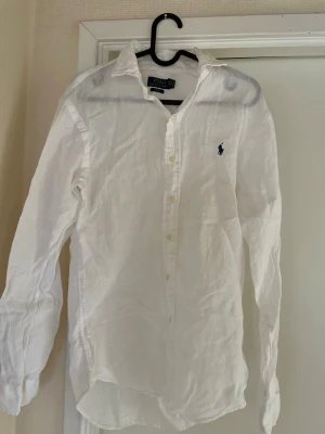 Vit linneskjorta från Polo Ralph Lauren - Klassisk vit skjorta från Polo Ralph Lauren i svalt linne. Skjortan har lång ärm, knappar framtill och den ikoniska broderade loggan på bröstet. Perfekt till jeans eller chinos för en clean och stilren look. Använd max 4 gånger inga tecken på användning 