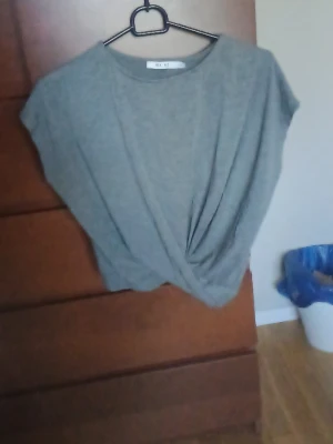 Grå croppad topp från NA-KD - Säljer en grå croppad topp från NA-KD med twistad detalj framtill. Toppen har korta ärmar och en avslappnad passform, perfekt till jeans eller kjol. Materialet känns mjukt och skönt mot huden.