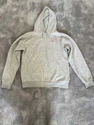 Grå hoodie med tryck 'JE M'EN AMOUR' från gina tricot - Grå hoodie med huva och trycket 'JE M'EN AMOUR' i rosa och svart på bröstet och större tryck på ryggen. Från gina tricot. Mjuk bomullsmix, ribbade muddar och avslappnad passform. Perfekt för en chill och trendig look.