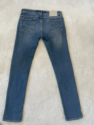 Jacob Cohen Jeans  - Säljer ett par blå slim fit jeans från Jacob Cohen med klassisk femficksdesign och snygga slitningar framtill. Jeansen har normal midja, tidlös passform och är tillverkade i mjuk denim med stretch för extra komfort. Perfekta för en avslappnad och trendig look.