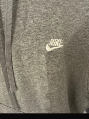 Grå hoodie från Nike med logga - Klassisk grå hoodie från Nike med vit broderad logga på bröstet. Hoodien har justerbar dragsko i huvan och ribbade muddar vid ärmar och nederkant. Tillverkad i mjuk bomullsblandning för maximal komfort.