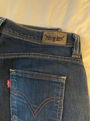 Mörkblå bootcut jeans från Levi's - Säljer ett par mörkblå jeans från Levi's med klassisk röd logga på bakfickan och bootcut-modell. Jeansen har fem fickor, lågmidjade och är tillverkade i slitstark denim. Midjemått: 40 cm, inrebensmått: 80 cm