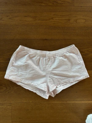 Ljusrosa shorts från Brandy Melville - Säljer ett par ljusrosa shorts från Brandy Melville med diskret randigt mönster. Shortsen har elastisk midja och två knappar framtill. Perfekta för sommardagar och riktigt sköna att bära.