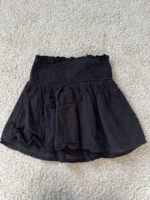 Svart kortkjol med smock - Säljer en svart tubtopp med volanger från Gina Tricot. 