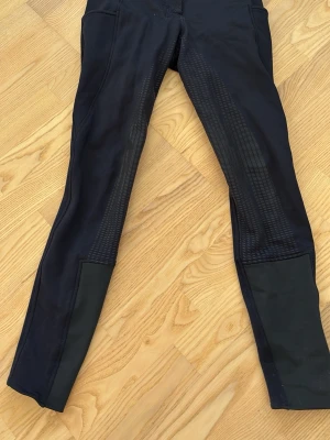 Svarta ridbyxor från Horze - Säljer ett par svarta ridbyxor från Horze med hög midja och två silverfärgade knappar framtill. Ridbyxorna har helskoning i silikon för extra grepp och stretchigt material som ger skön passform. De har även två fickor på båda sidor av låren. Perfekta för dig som vill ha både stil och funktion i stallet.