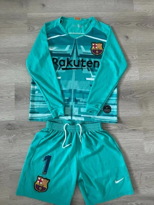 Barcelona målvaktströja och shorts - Säljer ett Barcelona målvaktsset med långärmad turkos tröja och matchande shorts. Tröjan har klubbmärke, Rakuten-logga och nummer 1 med Ter Stegen på ryggen. Shortsen har snörning och klubbmärke. Perfekt för fotbollsträning eller match. Storlek 152cm barn skick6/10 syns att den är använt men funkar ändå hur bra som helst kolla bilderna
