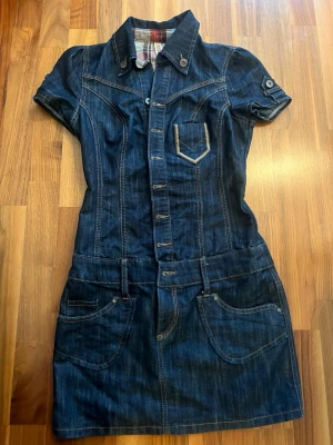 Mörkblå jeansklänning med knappar - Cool mörkblå jeansklänning med korta ärmar, krage och knappar hela vägen framtill. Klänningen har flera fickor och snygga sömdetaljer, perfekt för dig som gillar denim. Insidan av kragen har rutigt tyg för extra stil. storlek 32.