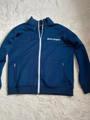 Blå zip-tröja från Palm Angels - Cool blå zip-tröja från Palm Angels med vit dragkedja och logga på bröstet. Tröjan har två fickor med dragkedja och hög krage. Perfekt för en avslappnad streetwear-look. Materialet är mjukt och stretchigt, vilket gör den bekväm att bära.