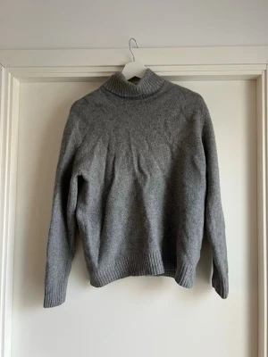 Grå stickad polotröja Calvin Klein - Mysig grå stickad polotröja från Calvin Klein med ribbade muddar och hög krage. Tröjan har en avslappnad passform och är perfekt för kyliga dagar. Materialet ser ut att vara ull eller en ullmix, vilket gör den både varm och skön. Barn storlek XXL motsvar vuxen storlek S / small. Måtten finns. 